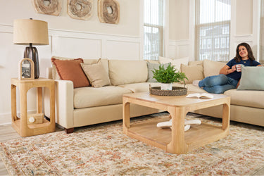 Cozy modern living room with beige sectional sofa, light wood coffee table and side table, patterned area rug, indoor potted plant, neutral decor and large windows with natural light, woman relaxing with coffee on sofa.