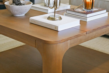 Mid-century modern solid wood coffee table with stacked home decor books, scented candle, and decorative bowl on a hardwood living room floor