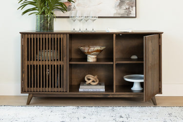 Mid-century modern wooden credenza with slatted doors, open shelves with a decorative bowl, books, plant and wine glasses.