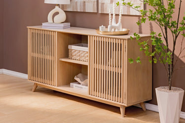 Mid-century modern natural oak sideboard with slatted doors and open shelving, styled with a woven basket, decorative lamp, and potted plant in a living room