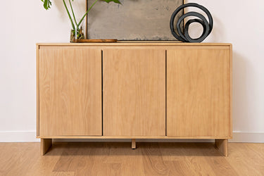 Light oak three-door sideboard cabinet (entryway console) with natural wood grain on hardwood floor, styled with potted plant and black circular sculpture.