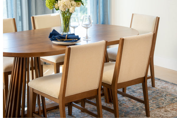 Round mid-century modern wooden dining table with beige upholstered chairs, floral centerpiece, wine glasses and navy napkins in a bright dining room