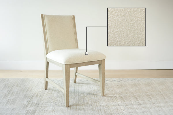 Beige upholstered dining chair with light oak legs and textured cream fabric, shown on a neutral rug against a white wall
