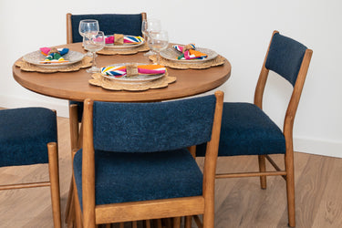 Mid-century modern round wooden dining table set with four navy upholstered chairs, woven placemats, colorful geometric napkins and stemmed wine glasses on hardwood floor