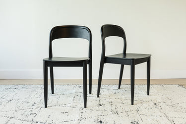 Pair of black modern wooden dining chairs on a textured white patterned rug against a white wall