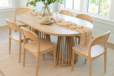 Light wood oval dining table with slatted pedestal base and six matching mid-century modern dining chairs, styled with a neutral table runner, glass vase and tableware in a bright coastal dining room