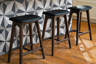 Three modern wooden bar stools with black upholstered seats at a kitchen island featuring geometric tile backsplash and hardwood floor.