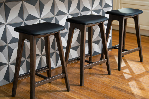 Three modern wooden bar stools with black upholstered seats at a kitchen island featuring geometric tile backsplash and hardwood floor.