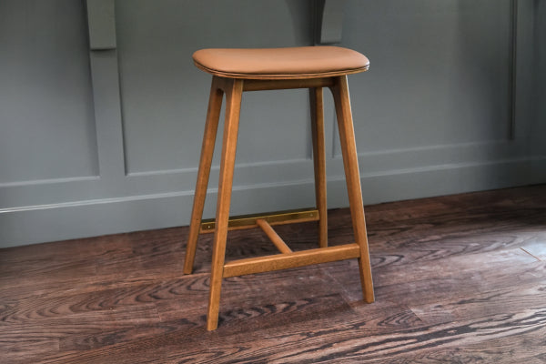 Mid-century modern wooden counter stool with tan leather seat on dark hardwood floor against gray paneled wall
