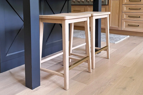Two light oak backless counter stools with brass footrest tucked under black kitchen island on wide-plank oak floors in a modern farmhouse kitchen