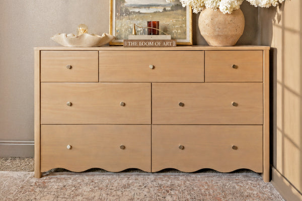Light natural wood seven-drawer dresser with brass knobs styled with a large ceramic vase of white flowers, framed landscape art and decorative bowl on top, sitting on a patterned rug against a neutral wall—bedroom furniture and modern farmhouse decor.