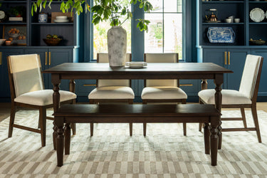 Sunlit solid wood dining table set with upholstered beige chairs and matching bench in a modern farmhouse dining room featuring navy built-in cabinets, a large ceramic vase centerpiece and neutral geometric rug.
