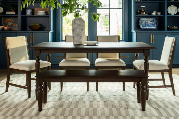 Sunlit solid wood dining table set with upholstered beige chairs and matching bench in a modern farmhouse dining room featuring navy built-in cabinets, a large ceramic vase centerpiece and neutral geometric rug.