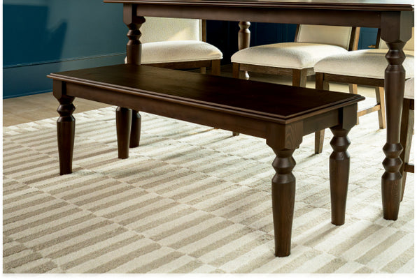 Dark wood dining bench with turned legs beneath matching farmhouse dining table and upholstered chairs on beige striped area rug