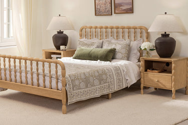Light-filled farmhouse bedroom with light-wood spindle bed, neutral patterned bedding, green lumbar pillow, matching wood nightstands and dark ceramic lamps