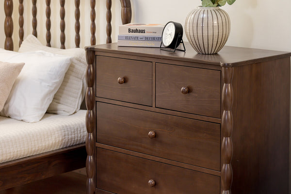 Dark walnut bedside dresser with four drawers and turned spindle bedposts, styled with stacked books, a black alarm clock and striped ceramic vase