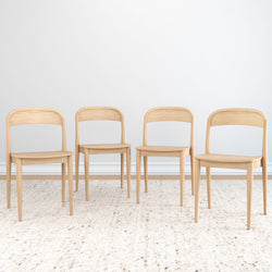 Set of four light natural wood dining chairs with curved backs on a textured neutral rug against a white wall, minimalist Scandinavian style.