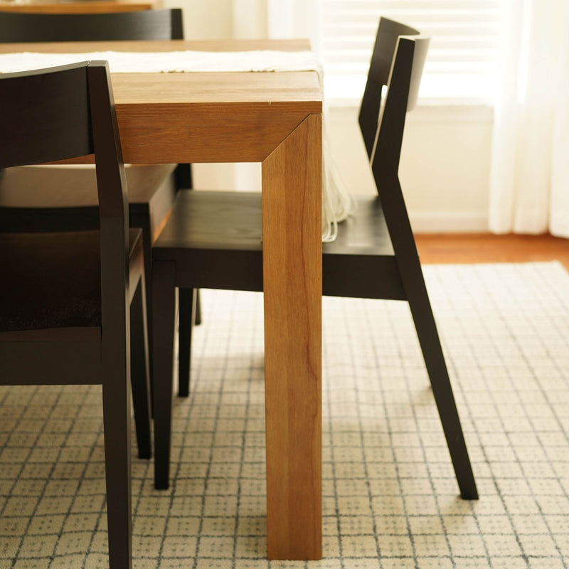 Close-up of a solid oak dining table leg with black modern dining chairs on a light patterned area rug in a bright minimalist dining room