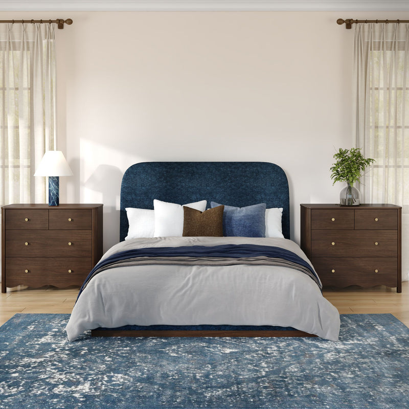 Modern bedroom with navy upholstered headboard, layered neutral bedding, wooden nightstands with lamps, sheer curtains, blue area rug and potted plant
