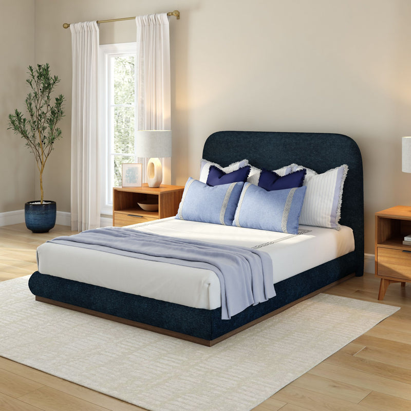Modern bedroom interior with navy upholstered bed, layered blue pillows, linen bedding, wooden nightstand and lamp, sheer white curtains