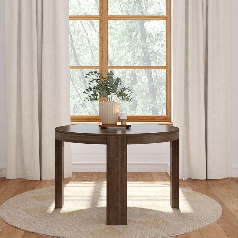 Round wooden table in a bright living room with a potted plant centerpiece, beige curtains, and hardwood flooring.