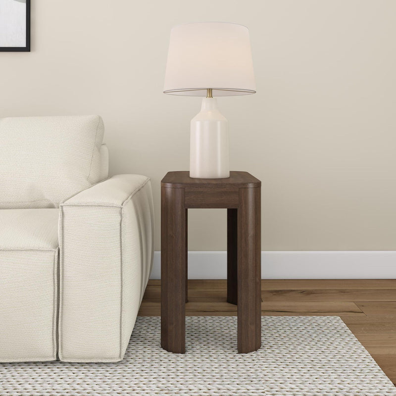 Wooden end table with white ceramic lamp next to cream-colored sofa in living room.