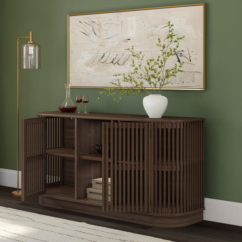 Dark wood slatted sideboard with open shelving and stacked books against a green accent wall, topped with abstract framed art, a white ceramic vase with leafy branches, a wine decanter and two glasses, and a brass floor lamp — modern mid-century living room decor.