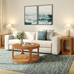 Modern coastal living room with cream sectional sofa, round wooden coffee table, teal patterned rug, abstract ocean wall art, wooden side tables, table lamps, and decorative pillows