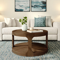 Round wooden coffee table styled with vase, candle and books in a modern living room with beige sofa, blue patterned pillow, abstract wall art and textured rug