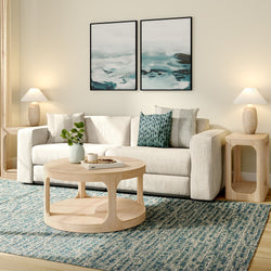 Modern coastal living room with beige fabric sofa, round light-wood coffee table, blue textured area rug, abstract ocean diptych wall art, patterned throw pillows, table lamps and potted plant