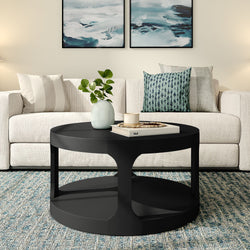 Black round coffee table with lower shelf styled with vase, candle and book in a modern neutral living room featuring a beige sofa, patterned throw pillows and blue area rug