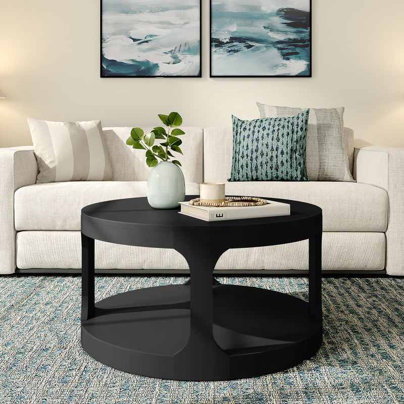 Black round coffee table with lower shelf styled with vase, candle and book in a modern neutral living room featuring a beige sofa, patterned throw pillows and blue area rug