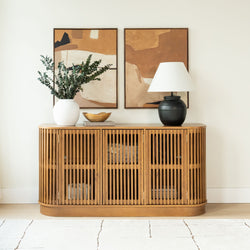 Wooden slatted console cabinet with white vase of eucalyptus, black ceramic lamp, gold bowl and abstract neutral wall art in a modern living room