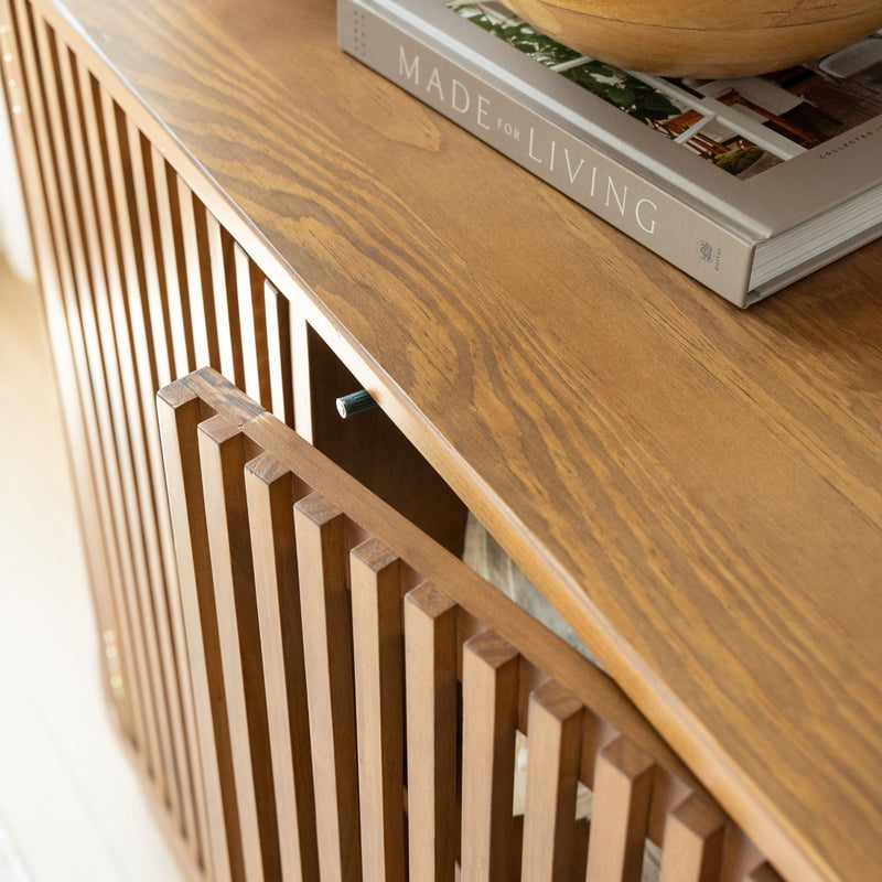 Close-up of mid-century modern slatted oak media console with curved top, decorative wooden bowl and "Made for Living" coffee table book — living room decor.