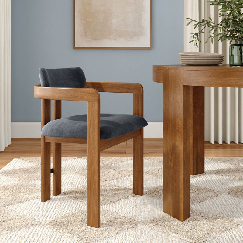 Minimalist modern dining room close-up showing an oak dining table and mid-century wooden chair with blue upholstered seat, textured jute rug, abstract wall art, stacked wooden plates and green glass vase