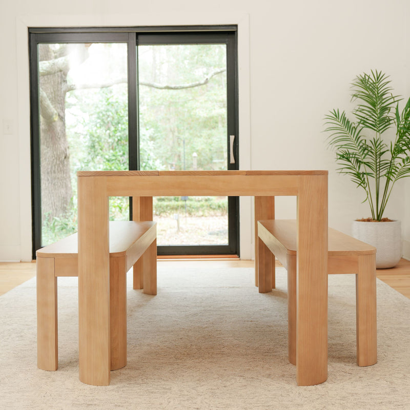 Minimalist natural wood dining table set with benches in modern bright room featuring large windows and indoor plant.