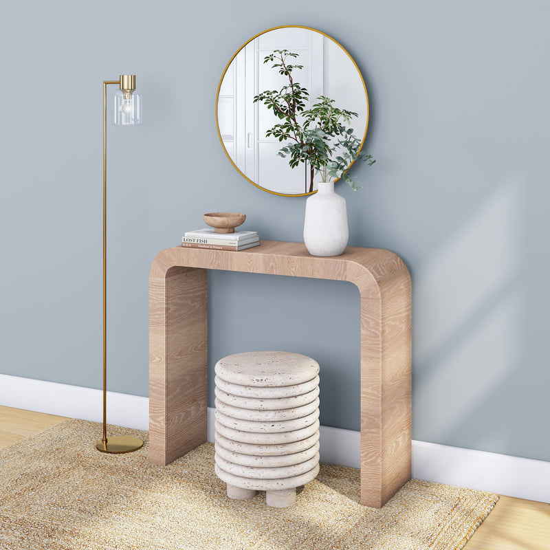 Minimalist entryway with light-wood curved console table against a powder blue wall, round gold-framed mirror, white ceramic vase with greenery, gold floor lamp and textured natural rug