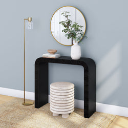 Modern minimalist entryway with black arched console table, round gold mirror, white ceramic vase with greenery, stacked stone stool and brass floor lamp on woven rug.