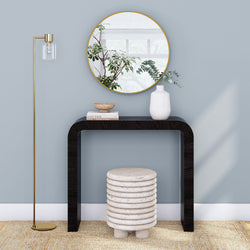 Modern entryway console table vignette — black curved console with round gold mirror, white ceramic vase with eucalyptus, stacked stone stool, brass floor lamp and neutral rug on light blue wall, minimalist home decor interior
