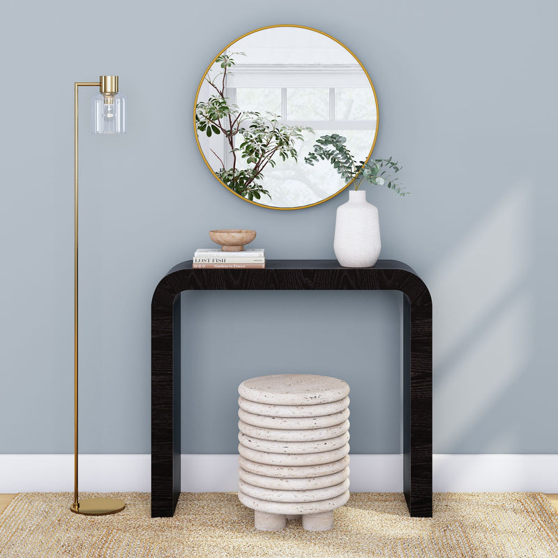 Modern entryway console table vignette — black curved console with round gold mirror, white ceramic vase with eucalyptus, stacked stone stool, brass floor lamp and neutral rug on light blue wall, minimalist home decor interior