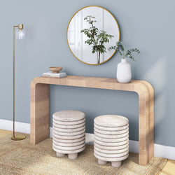 Minimalist modern entryway: light wood curved console table beneath a round gold-framed mirror, brass floor lamp, white ceramic vase with green foliage, and two stacked ribbed stone stools on a textured rug against a soft blue wall.