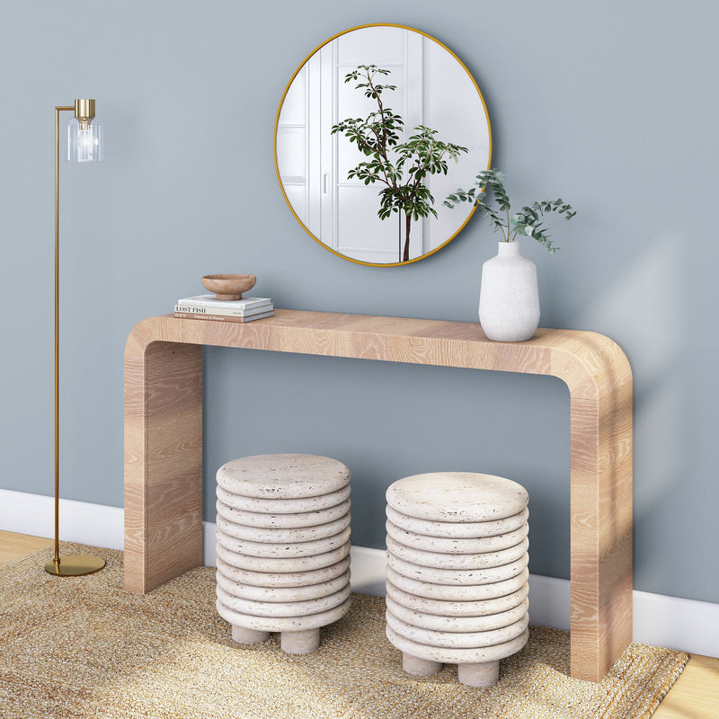 Minimalist modern entryway: light wood curved console table beneath a round gold-framed mirror, brass floor lamp, white ceramic vase with green foliage, and two stacked ribbed stone stools on a textured rug against a soft blue wall.