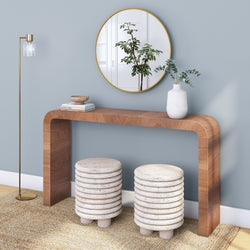 Minimalist modern entryway with curved wood console table, gold-framed round mirror, white ceramic vase with eucalyptus, stacked ribbed stone stools on textured rug and brass floor lamp against a blue wall