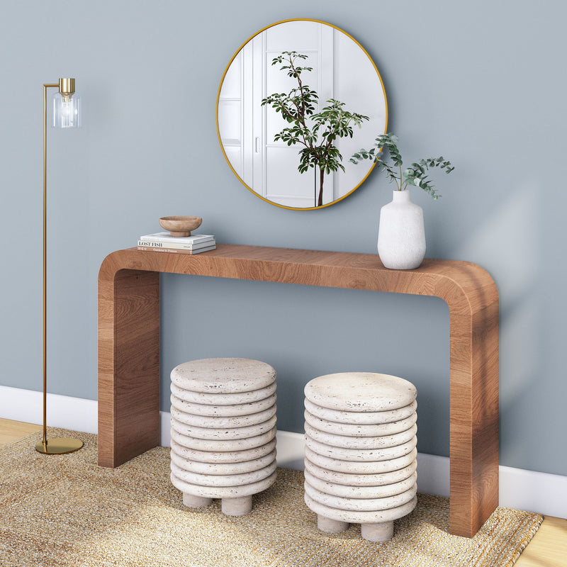 Minimalist modern entryway with curved wood console table, gold-framed round mirror, white ceramic vase with eucalyptus, stacked ribbed stone stools on textured rug and brass floor lamp against a blue wall