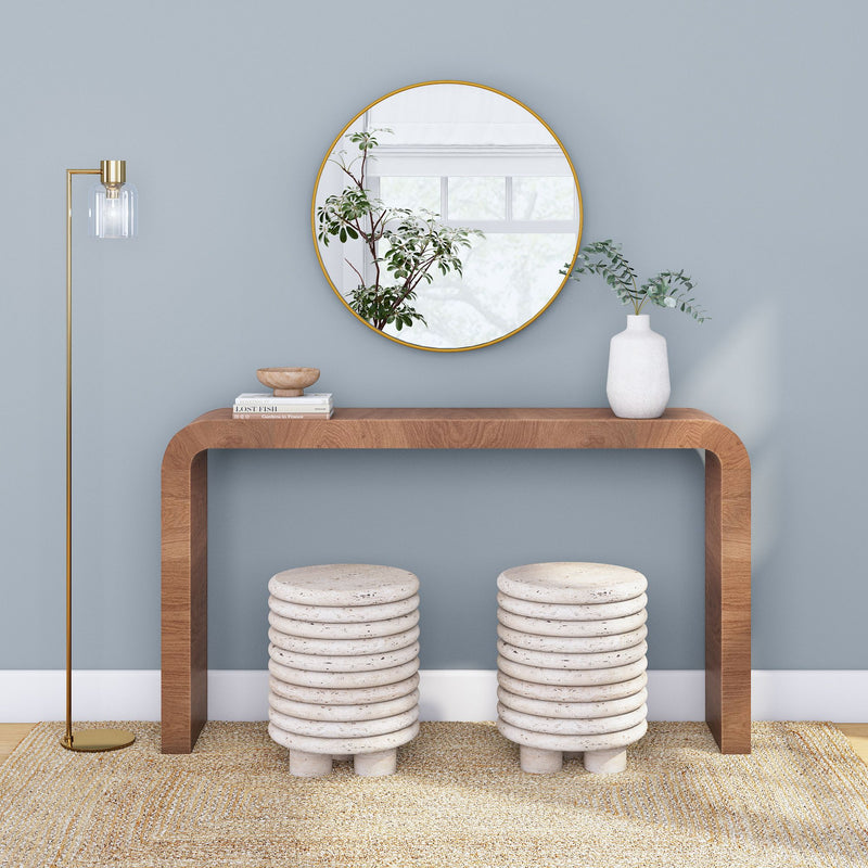 Modern minimalist entryway: curved walnut console table beneath round gold mirror, white ceramic vase with eucalyptus, two stacked stone stools, brass floor lamp on blue-gray wall with natural jute rug