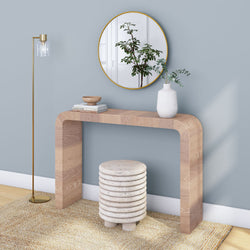 Modern entryway wood console table with round gold mirror, white vase with eucalyptus, stacked stone stool, gold floor lamp and neutral rug on blue-gray wall