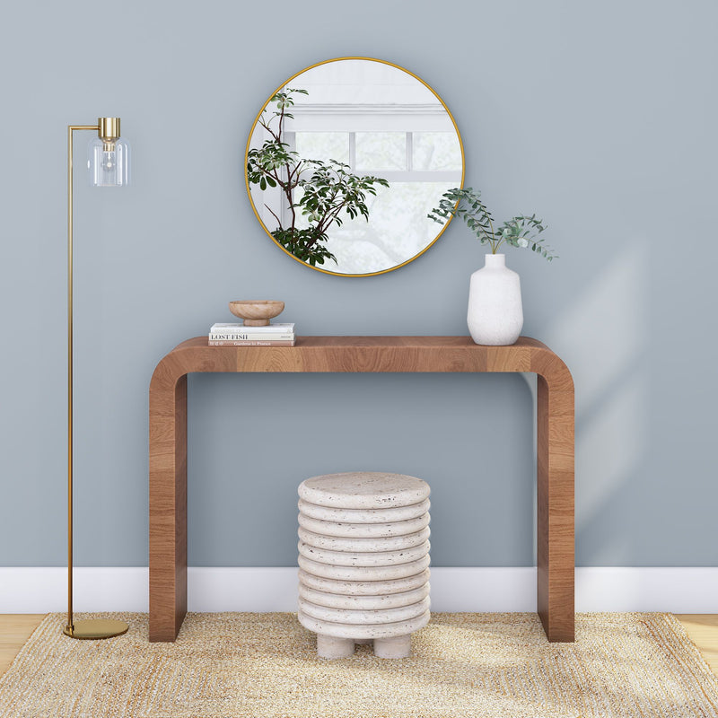 Minimalist entryway vignette with curved wooden console table, round gold-framed mirror reflecting a window and plant, white ceramic vase with eucalyptus, stacked ribbed stone stool, brass floor lamp, light blue wall and natural jute rug
