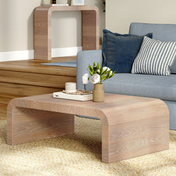 Light oak curved modern coffee table in a Scandinavian living room with a blue sofa, striped and navy cushions, vase of white flowers, book and candle on a woven rug