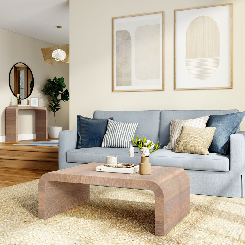 Modern minimalist living room with light-blue sofa, curved wooden coffee table, beige rug, abstract framed art, throw pillows and potted plant
