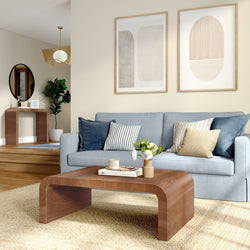 Modern living room interior with light blue sofa, striped and textured throw pillows, curved wooden coffee table with vase and books, neutral abstract wall art, indoor plant, natural woven rug and minimalist decor